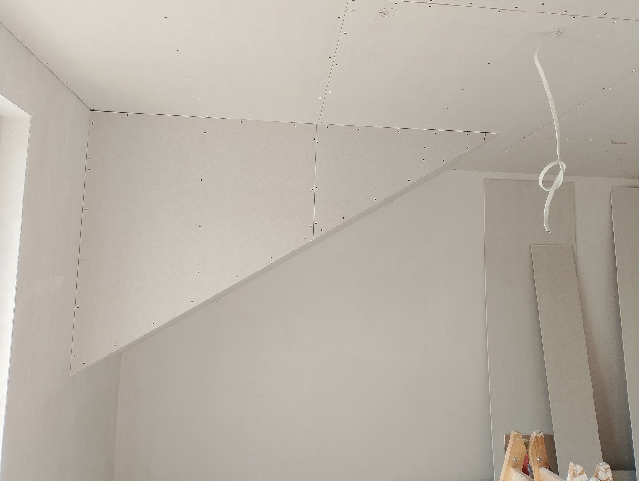 Skośny sufit i ściana pokryte płytami gipsowo-kartonowymi, widoczne łączenia i śruby, fragment okna z lewej strony, zwisający kabel i płyty w tle.
