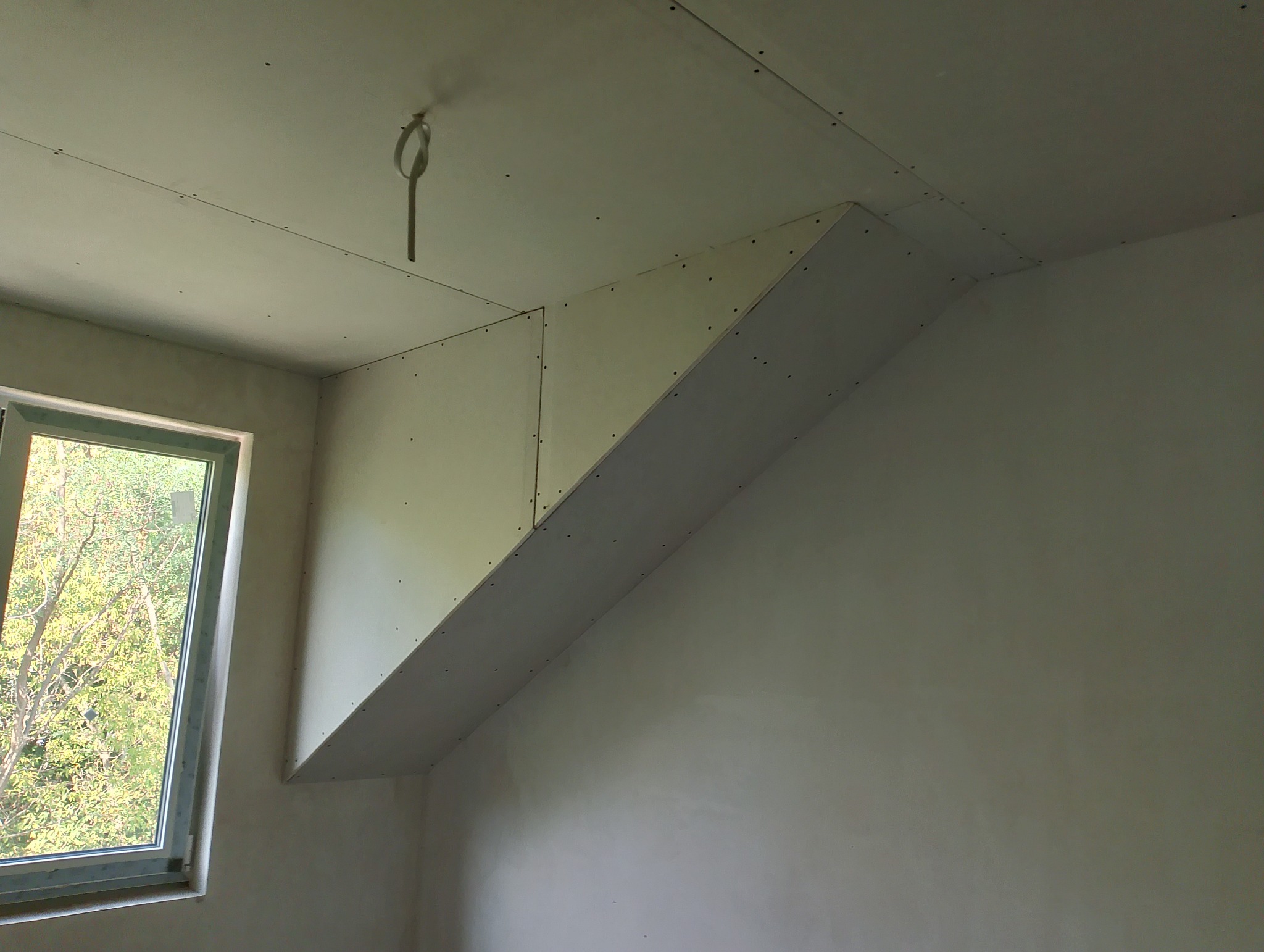 Skośny sufit poddasza z płyt kartonowo-gipsowych, widoczne łączenia płyt i wkręty, surowy stan przed szpachlowaniem i malowaniem, okno z widokiem na zieleń.