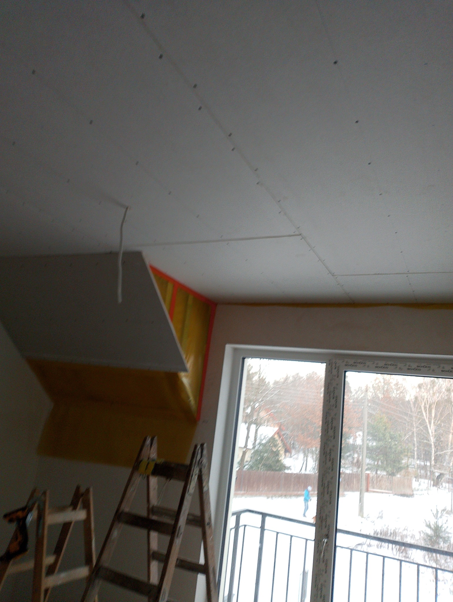 Montaż płyty gipsowo-kartonowej na poddaszu, widoczne rusztowanie, okno balkonowe z widokiem na zimowy krajobraz.