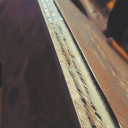 Dudush - Długi, potrójny spaw na krawędzi grubej, stalowej płyty, widoczny z bliska pod kątem, z wyraźnymi śladami po spawaniu.