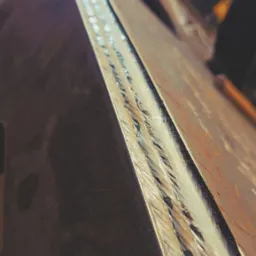 Długi, potrójny spaw na krawędzi grubej, stalowej płyty, widoczny z bliska pod kątem, z wyraźnymi śladami po spawaniu.