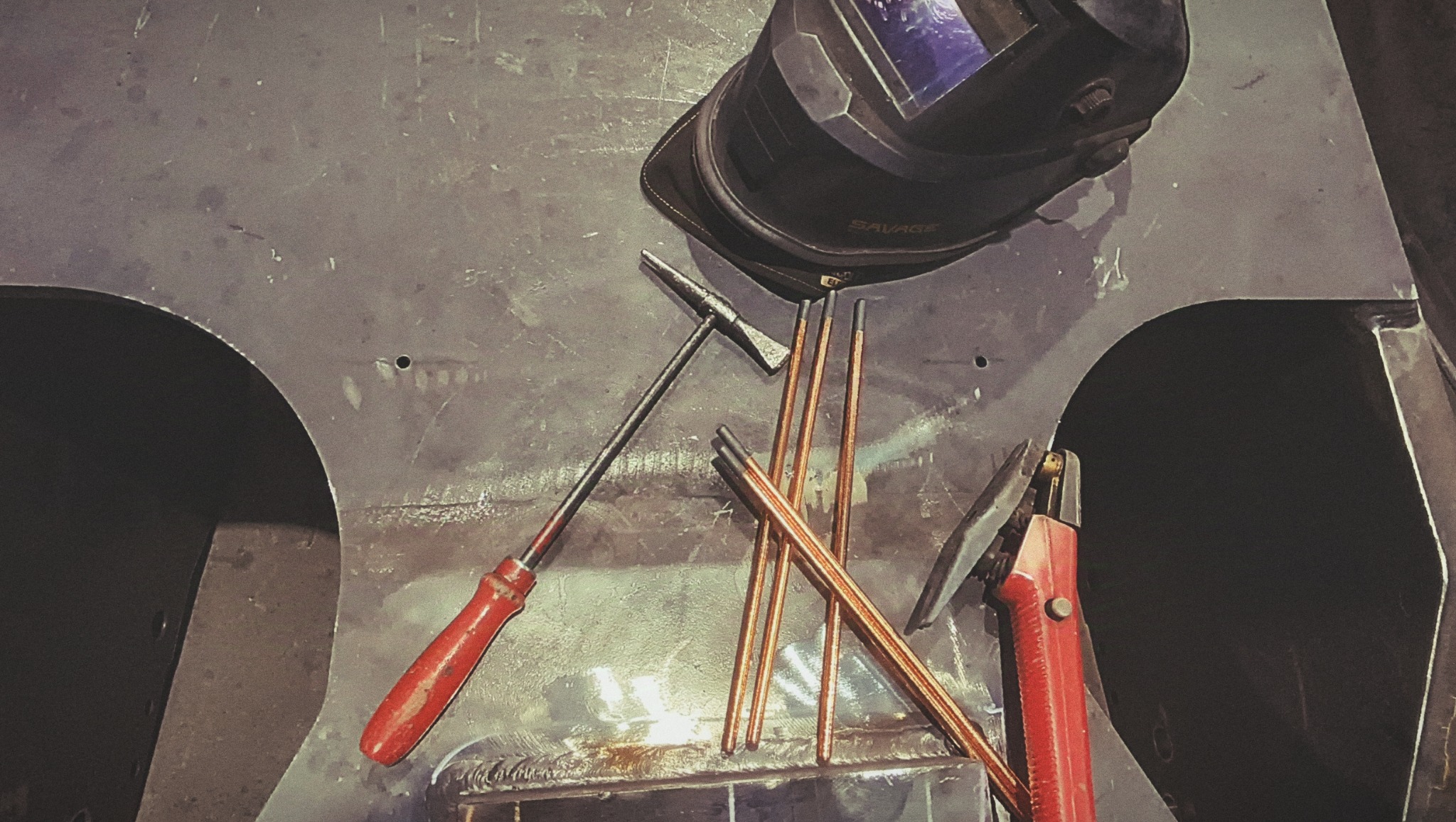 Maska spawalnicza, młotek, elektrody i uchwyt elektrody na metalowej powierzchni z widocznymi śladami spawania.