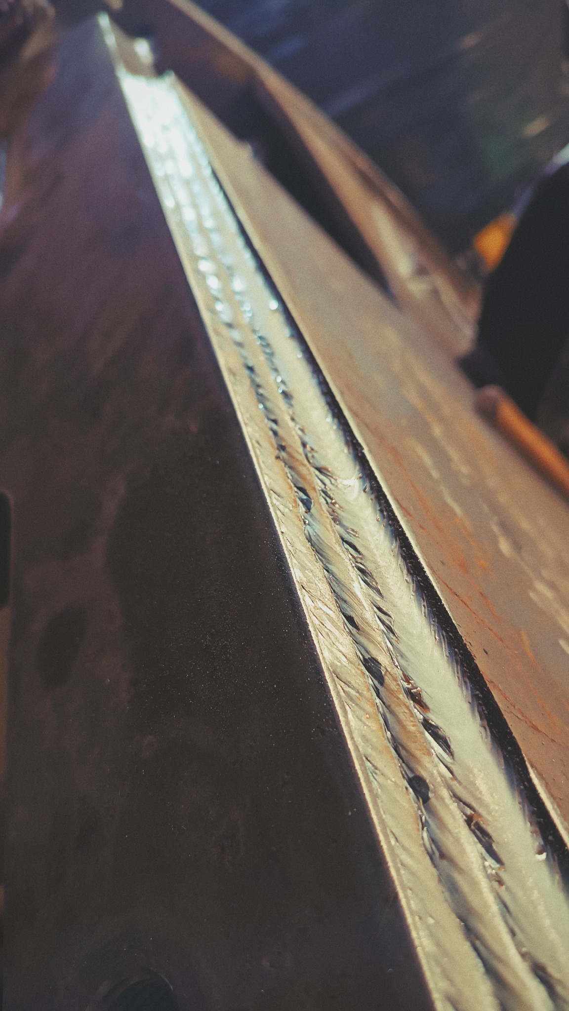 Długi, potrójny spaw na krawędzi grubej, stalowej płyty, widoczny z bliska pod kątem, z wyraźnymi śladami po spawaniu.