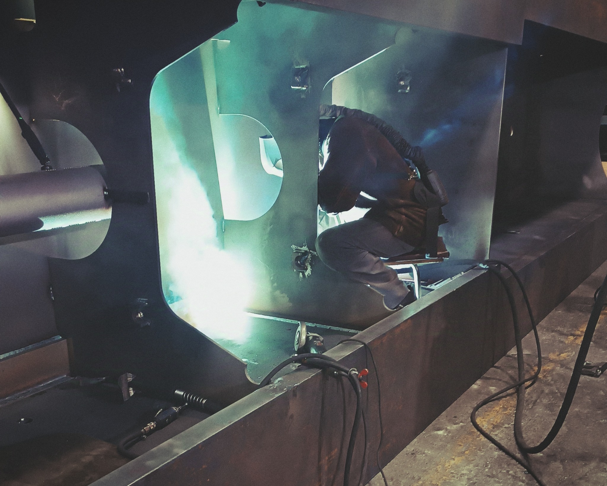Pracownik w kombinezonie spawalniczym, siedząc na niskim stołku, spawa element konstrukcyjny z grubych metalowych płyt, widoczne iskry i narzędzia spawalnicze.