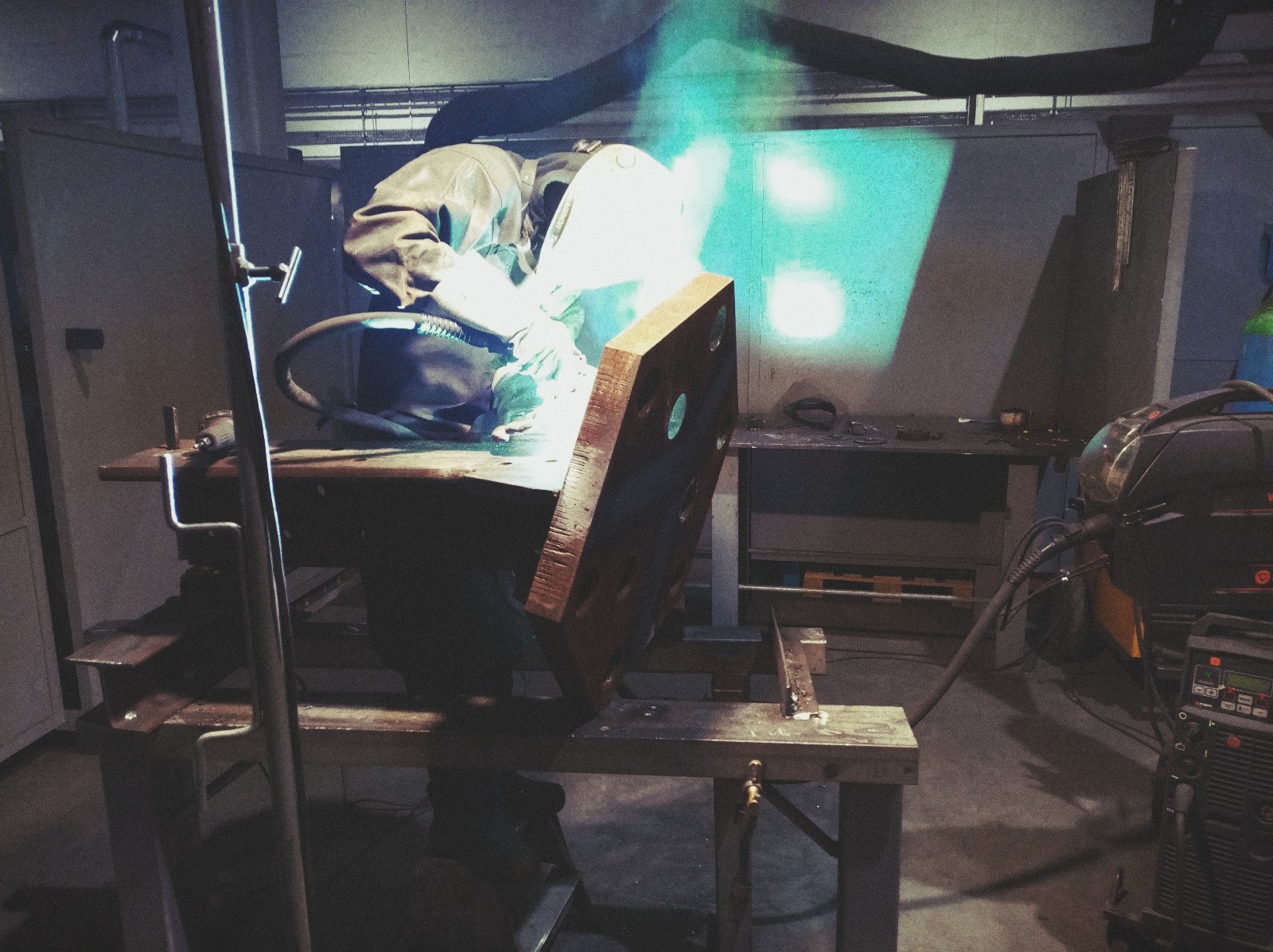 Pracownik w kombinezonie i masce ochronnej spawa gruby, drewniany element z otworami na metalowym stole warsztatowym; widoczny sprzęt spawalniczy i odciąg dymu.