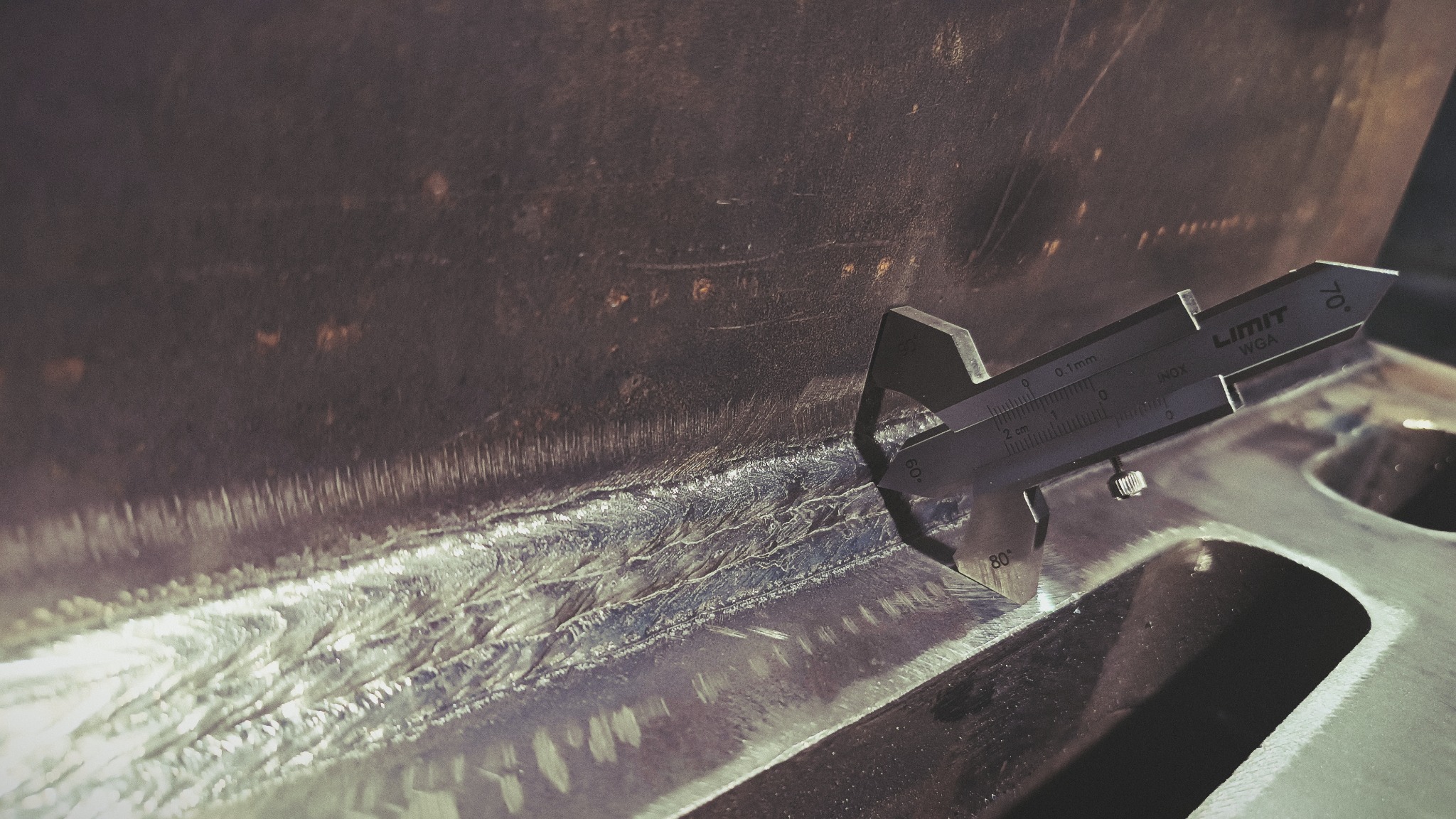 Pomiar spoiny spawalniczej suwmiarką 'LIMIT WGA' na metalowej konstrukcji z widocznymi śladami obróbki i wcześniejszych spawów.