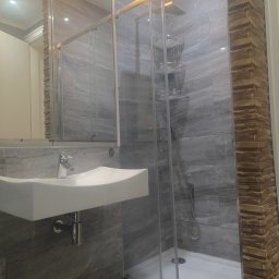 Ref con - Wykończona łazienka z kabiną prysznicową, umywalką i lustrem. Ściany wyłożone szarymi płytkami imitującymi kamień i elementami dekoracyjnymi z cegły.