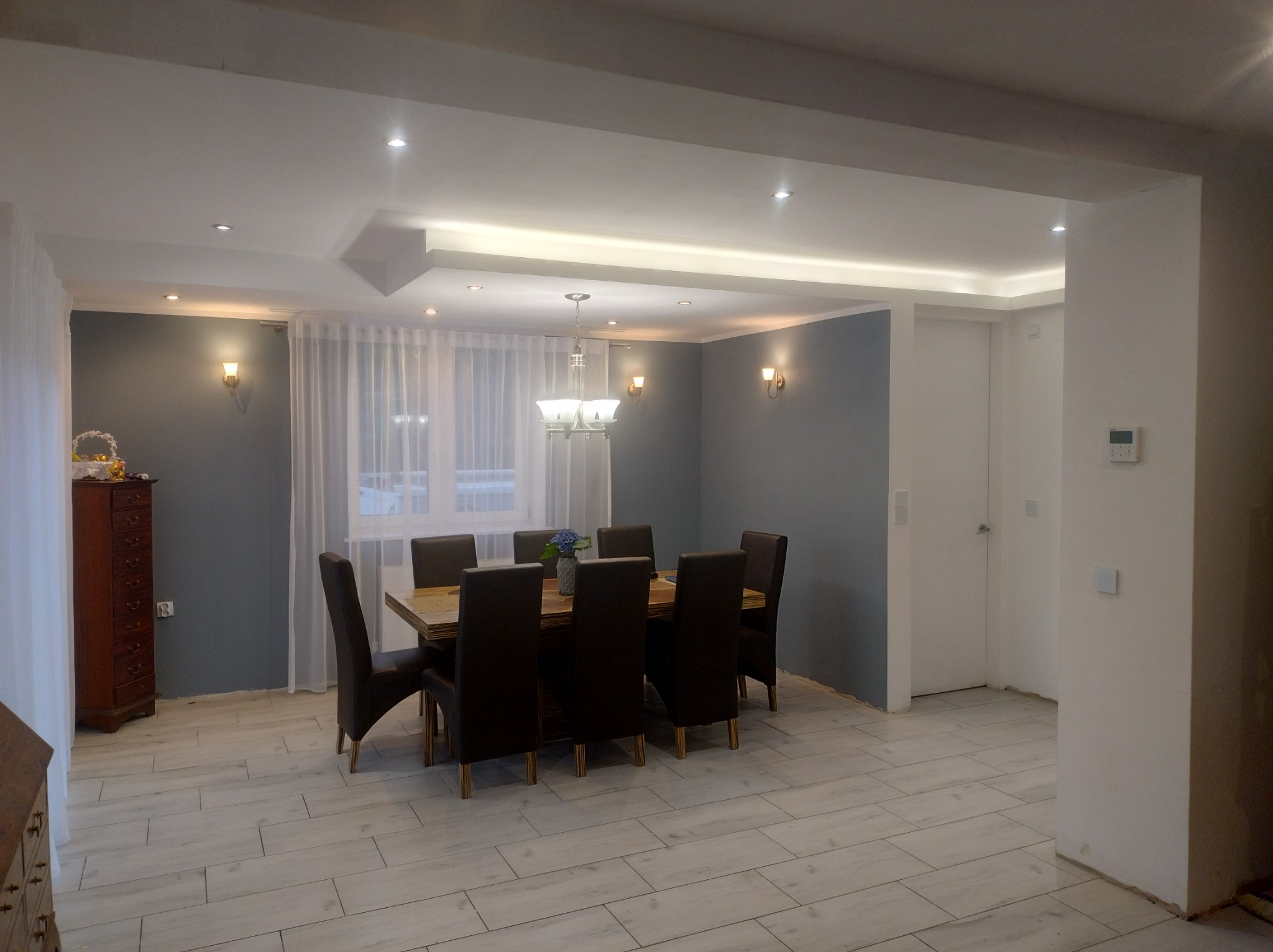 Jasne wnętrze jadalni z drewnianym stołem, krzesłami i subtelnym oświetleniem punktowym. Nowoczesny design podkreślają szare ściany i białe płytki podłogowe.