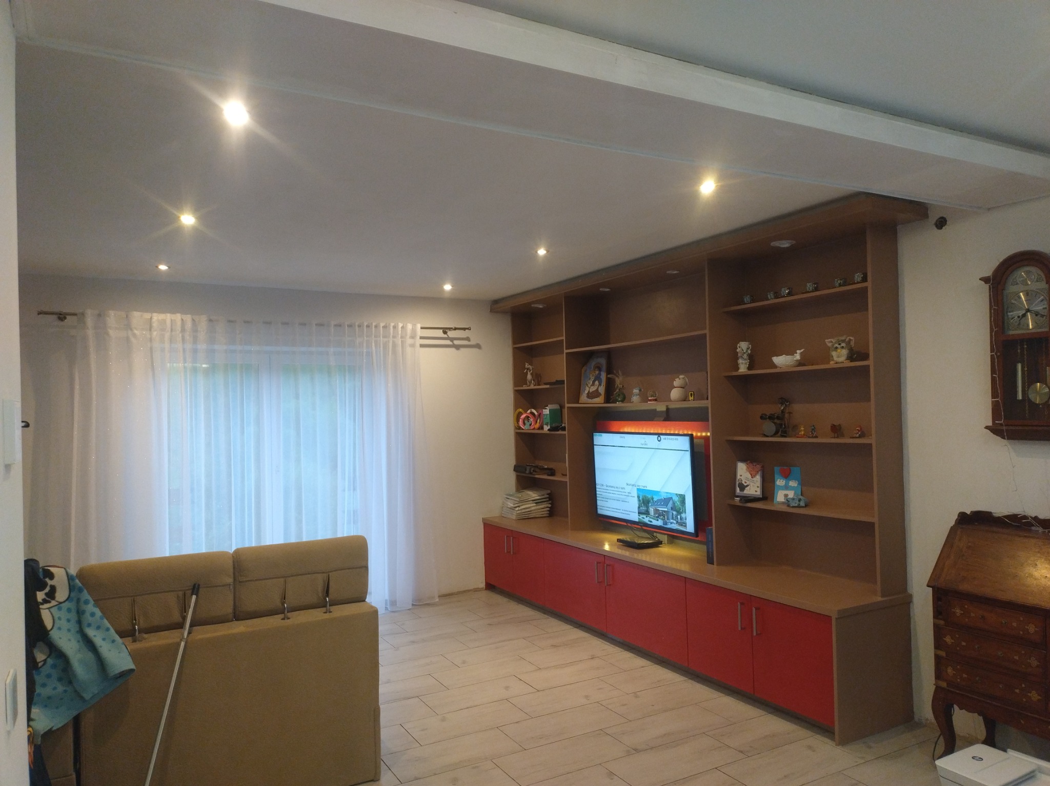 Jasne wnętrze salonu z beżową sofą i zabudową meblową z czerwonymi frontami. Na półkach dekoracje i książki. Podłoga wyłożona płytkami imitującymi drewno.