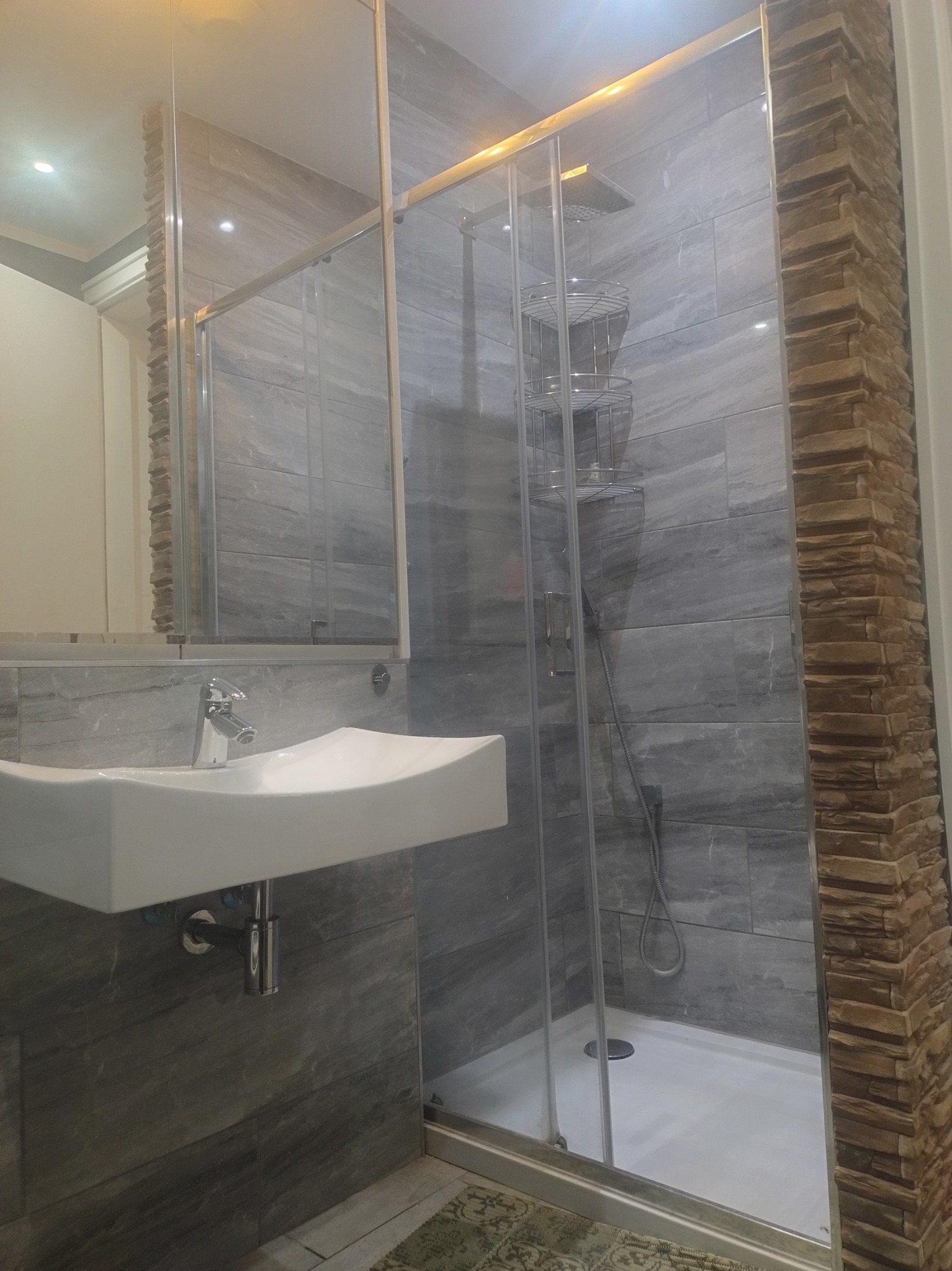 Wykończona łazienka z kabiną prysznicową, umywalką i lustrem. Ściany wyłożone szarymi płytkami imitującymi kamień i elementami dekoracyjnymi z cegły.