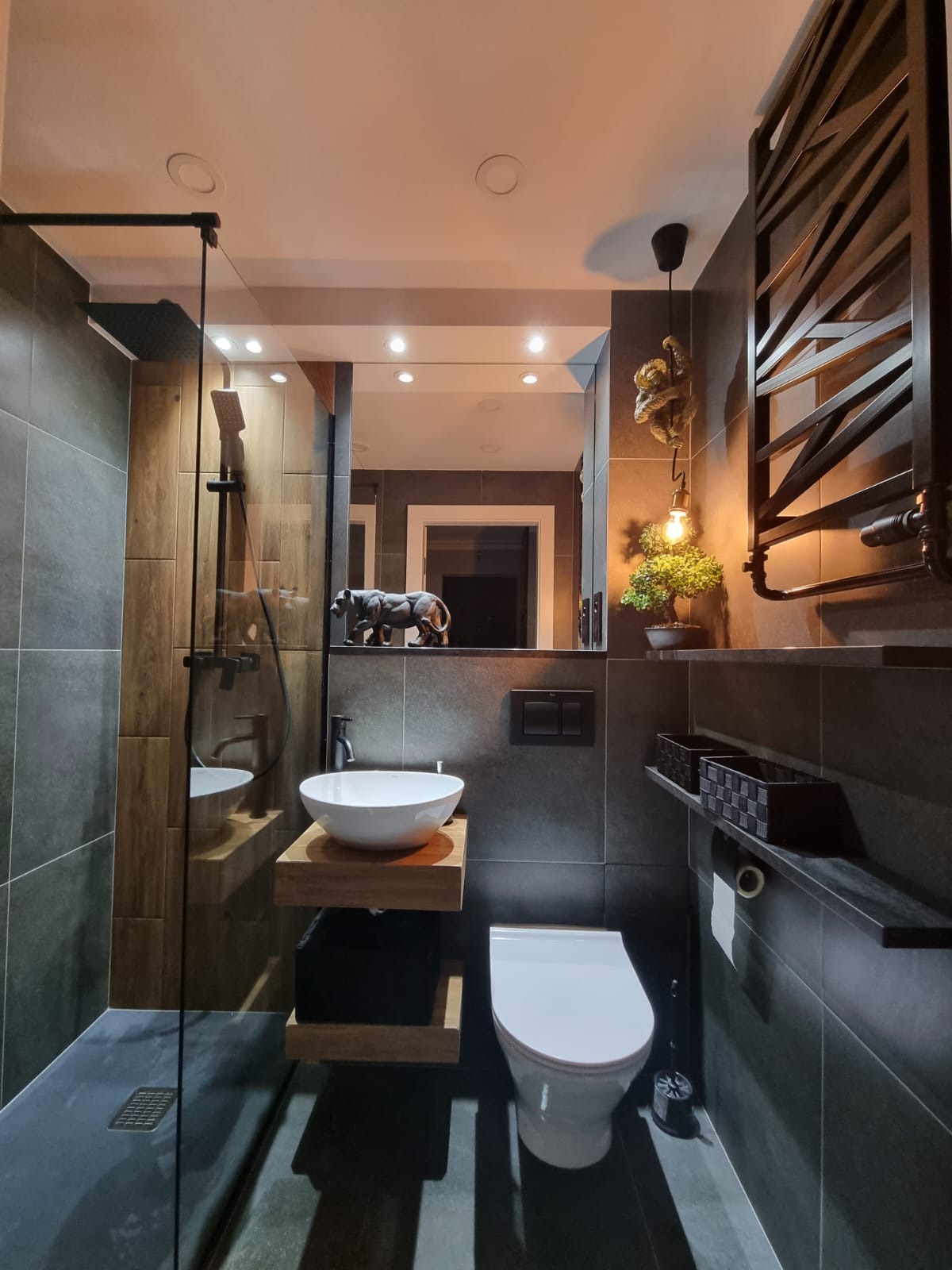 Nowoczesna łazienka z szarymi płytkami, przeszklonym prysznicem, umywalką nablatową na drewnianym blacie, wiszącą toaletą oraz dekoracjami: figurką zwierzęcia na lustrze i lampką z małpką.