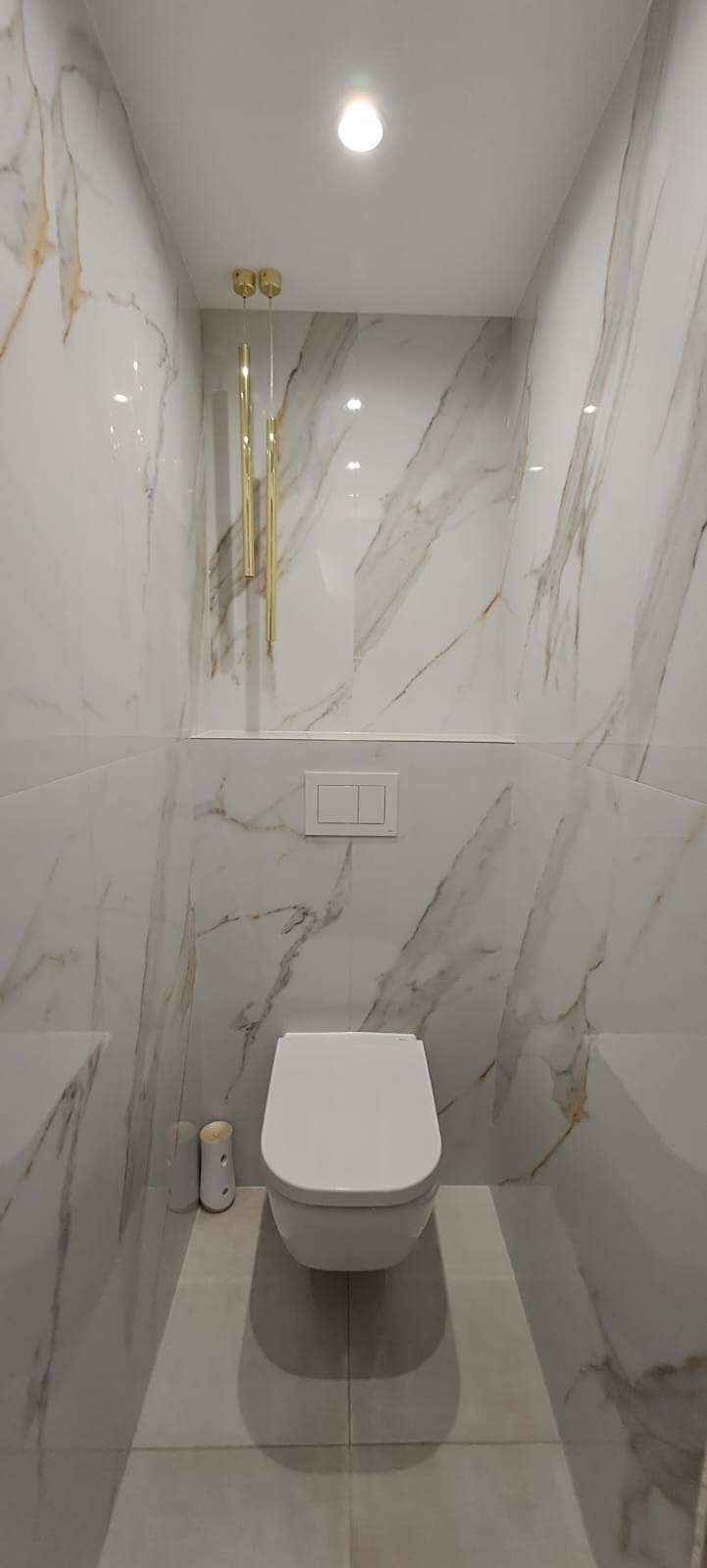 Zawieszona miska WC w nowoczesnej łazience wykończonej białymi płytkami imitującymi marmur z złotymi żyłami, podwieszany stelaż z dwudzielnym przyciskiem spłukującym, wiszące złote lampy tuby.