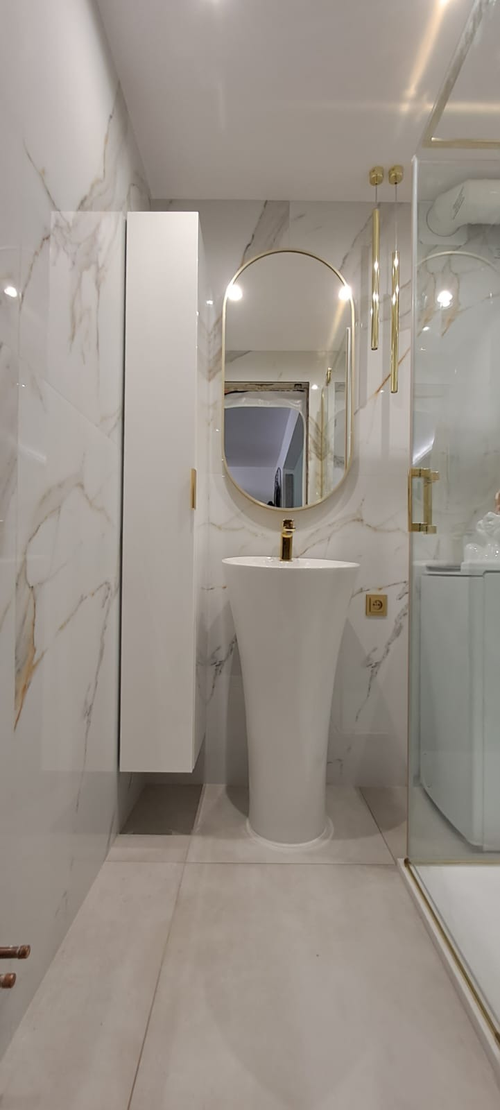 Elegancka łazienka z białą umywalką kolumnową, złotą armaturą i owalnym lustrem w złotej ramie, marmurowe płytki na ścianach, wisząca szafka i szklana kabina prysznicowa.