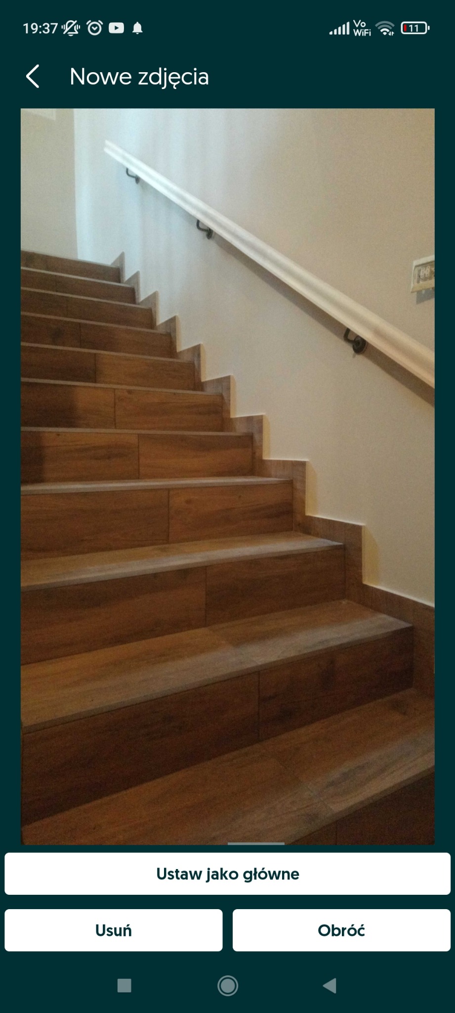 Schody wyłożone płytkami imitującymi drewno, z białą poręczą i szarą ścianą w tle.