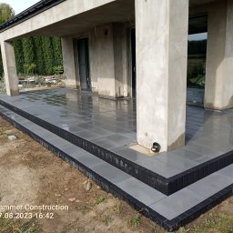 od a do z centrum dysyrybucji - Nowoczesny taras z szarych płyt, wykończony czarną kostką brukową, przylegający do surowego, betonowego budynku w otoczeniu zieleni.