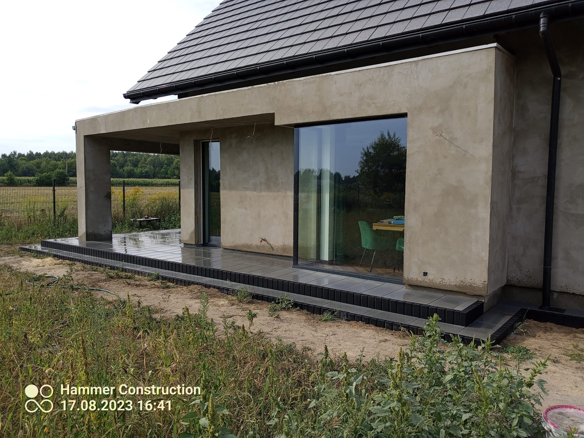 Surowy betonowy dom z nowoczesnym tarasem wyłożonym szarą kostką brukową, widok z poziomu gruntu na tle łąki.