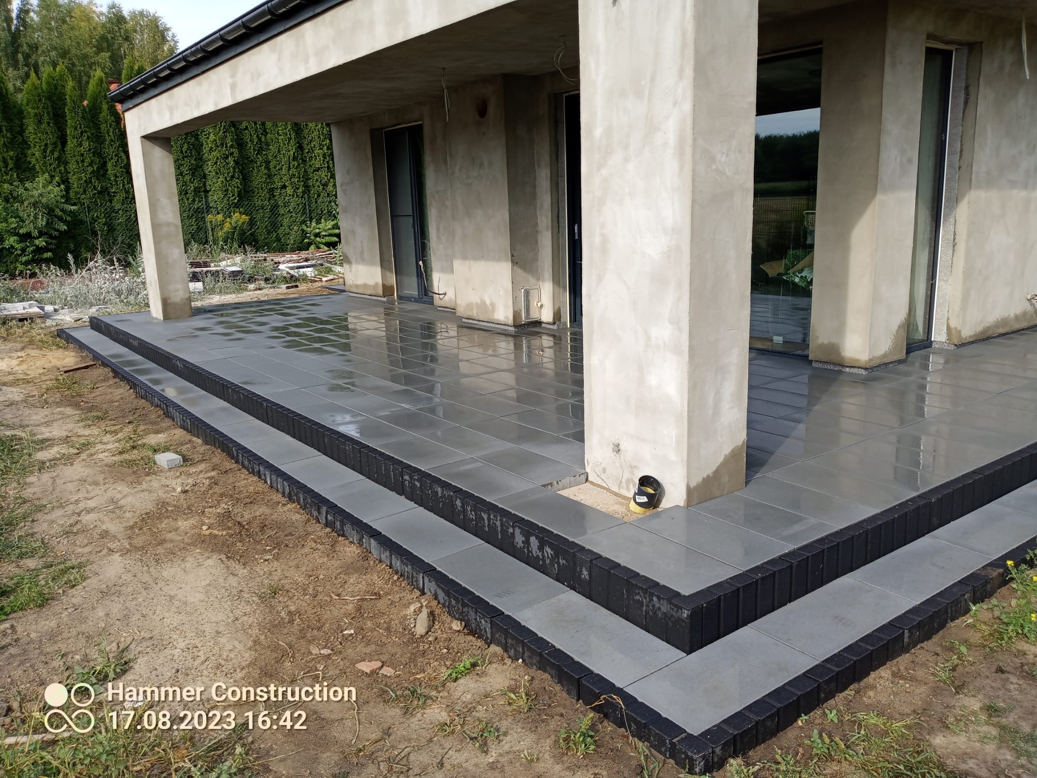 Nowoczesny taras z szarych płyt, wykończony czarną kostką brukową, przylegający do surowego, betonowego budynku w otoczeniu zieleni.