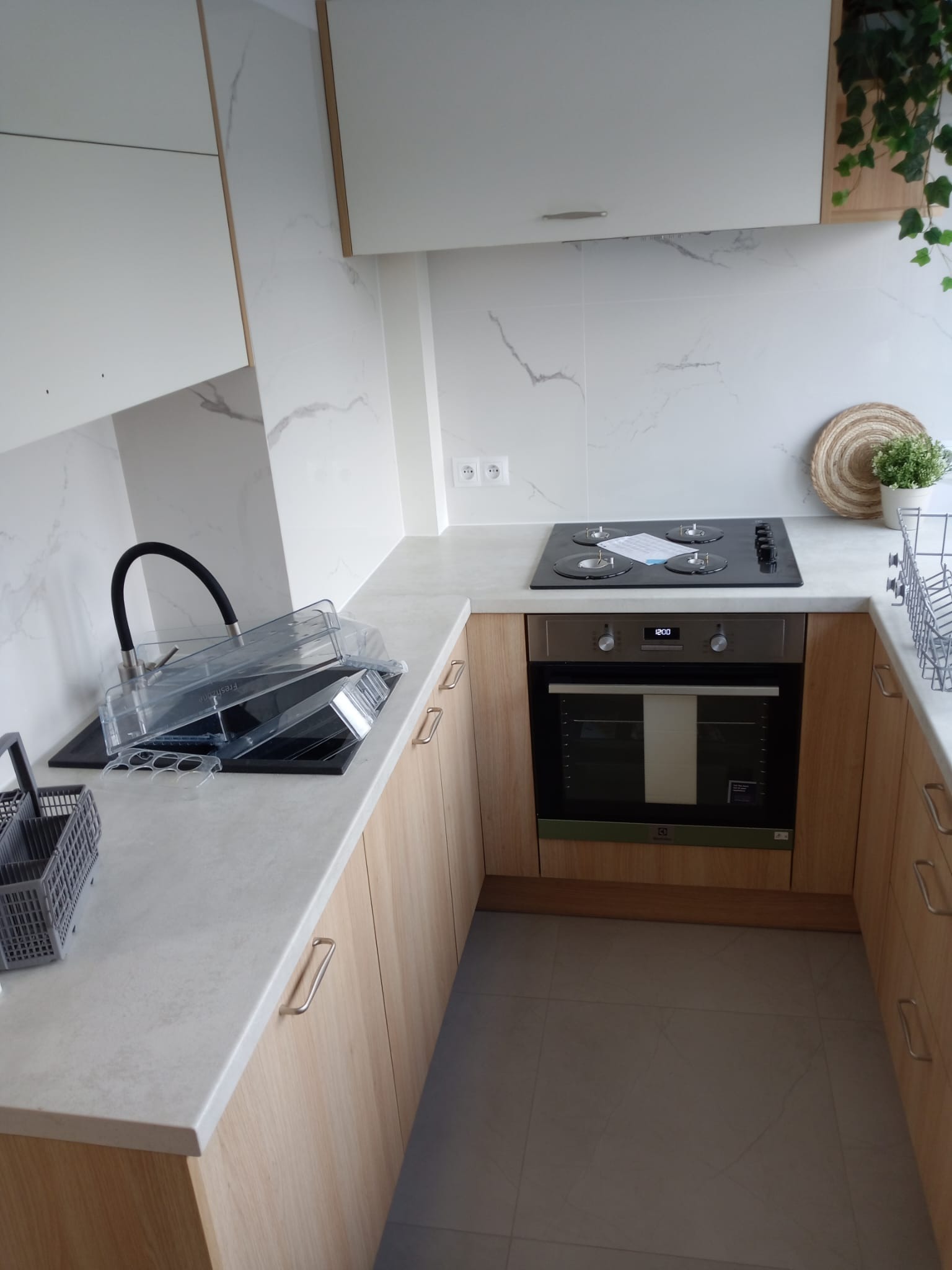 Nowoczesna kuchnia z zabudową w odcieniach bieli i drewna, wyposażona w płytę grzewczą, piekarnik, zlew z czarną armaturą i suszarką, oraz dekoracje roślinne na blacie.