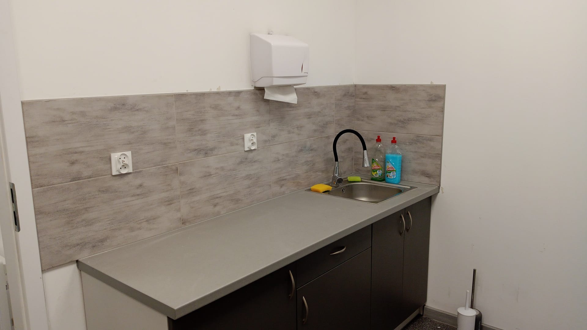 Wykończony aneks kuchenny z szarymi płytkami imitującymi drewno, blatem w kolorze popielatym i ciemnymi szafkami, wyposażony w zlewozmywak, baterię, dozownik ręczników papierowych i środki czystości.