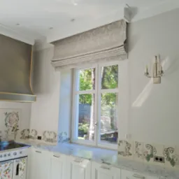 Beżowa, wzorzysta roleta w stylu rzymskim zamontowana w oknie eleganckiej kuchni z marmurowym blatem i dekoracyjnymi płytkami ceramicznymi.