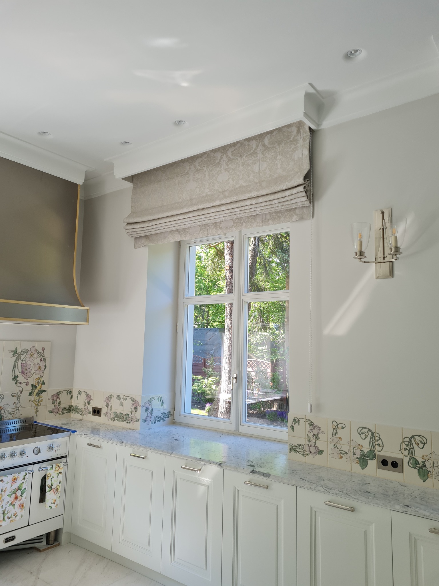 Beżowa, wzorzysta roleta w stylu rzymskim zamontowana w oknie eleganckiej kuchni z marmurowym blatem i dekoracyjnymi płytkami ceramicznymi.