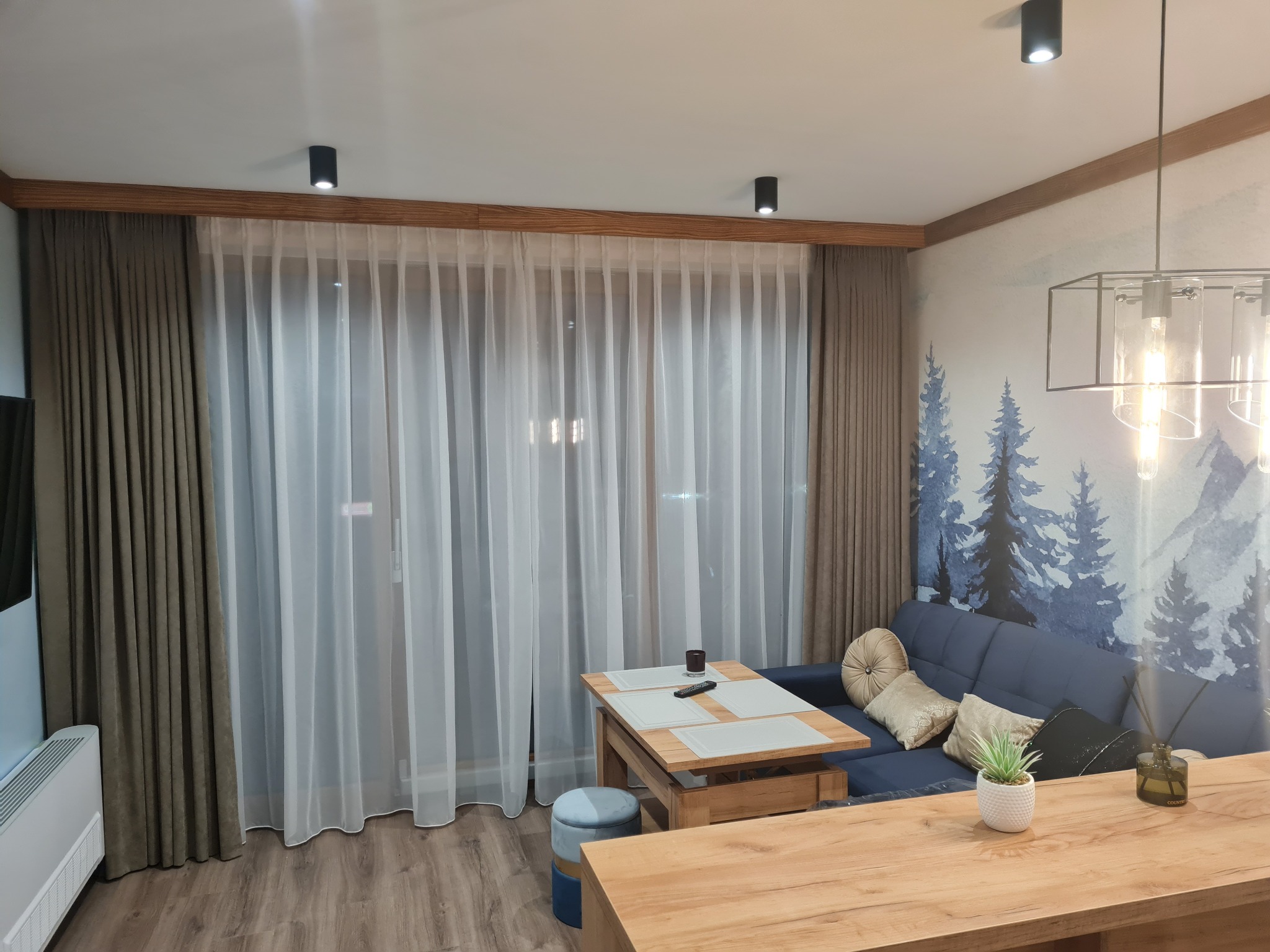 Jasne wnętrze salonu z drewnianymi akcentami, białymi zasłonami i szarymi kotarami, uzupełnione tapetą z motywem zimowego lasu i nowoczesnym oświetleniem sufitowym.
