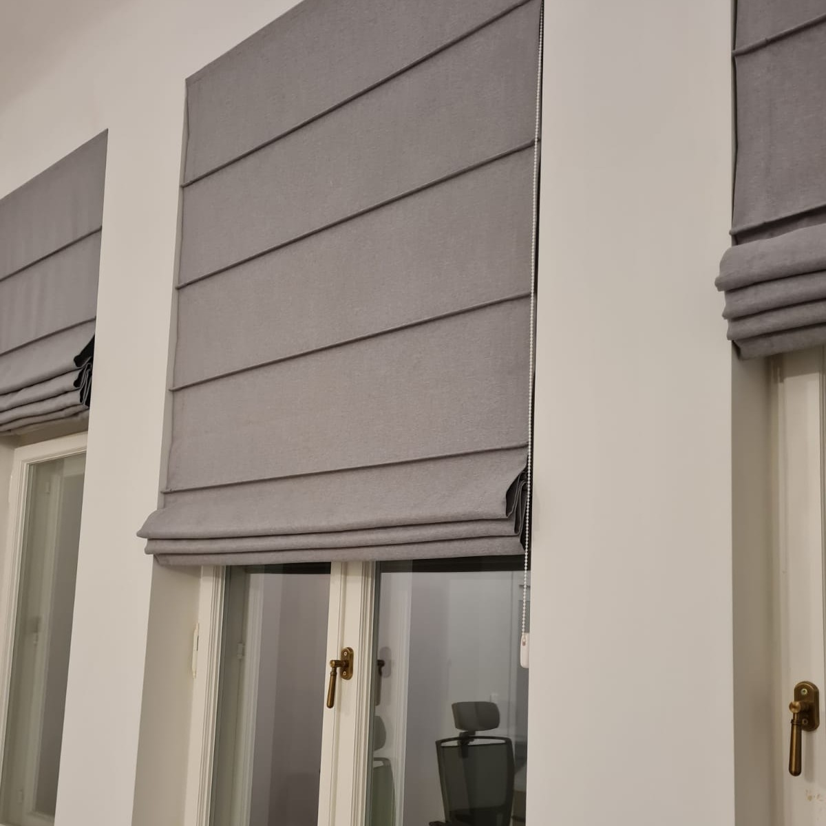 Szare, tekstylne rolety w stylu rzymskim zamontowane w oknach, widoczny mechanizm łańcuszkowy i fragment biurowego krzesła w tle.