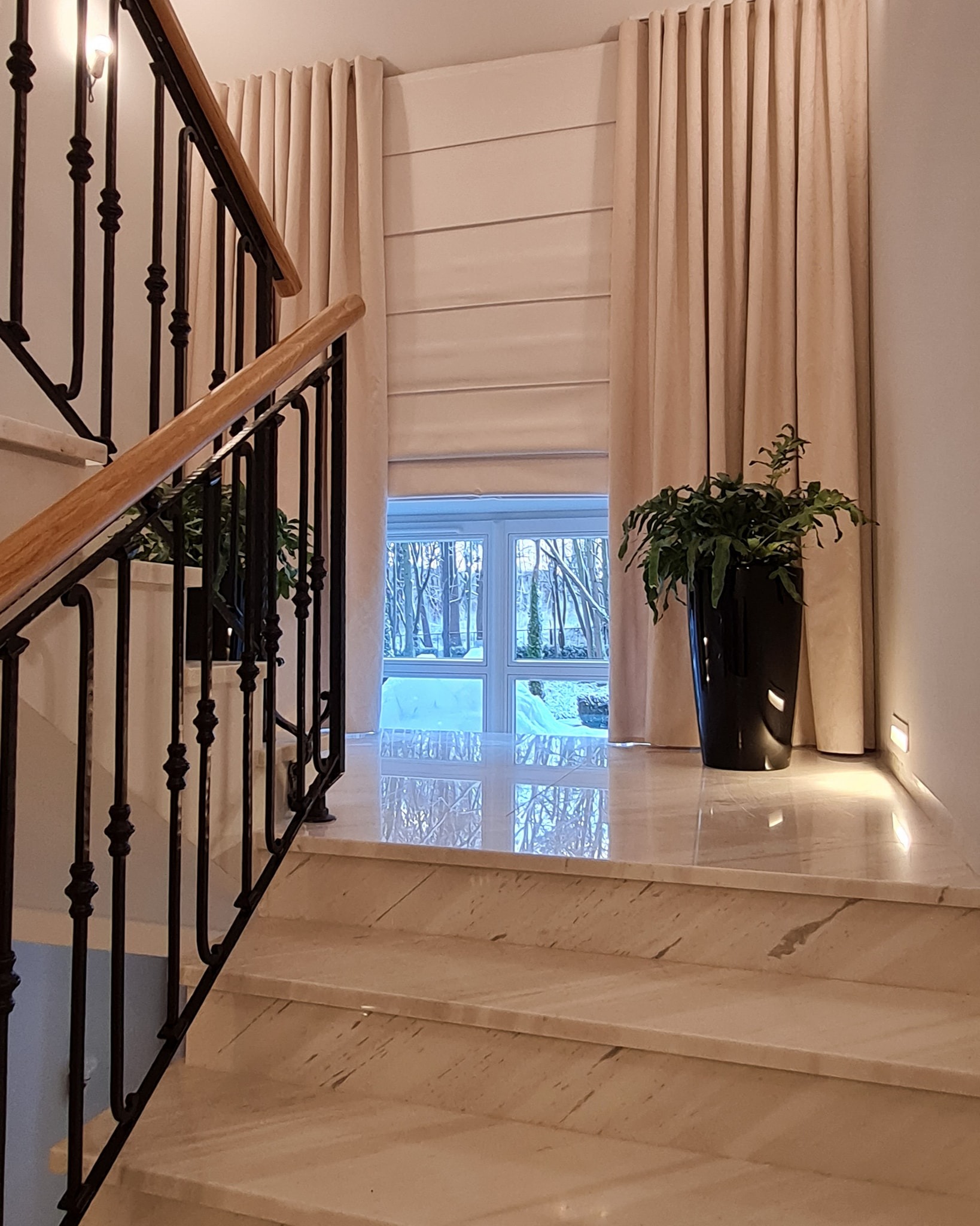 Eleganckie wnętrze z marmurowymi schodami, czarną balustradą, kremowymi zasłonami i roletą rzymską w tle, widok na zimowy ogród przez okno.