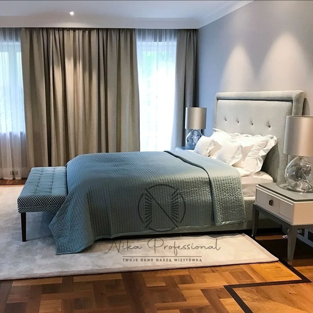 Elegancka sypialnia w odcieniach szarości i beżu z łóżkiem, pikowaną narzutą, szafkami nocnymi z lampkami i zasłonami do podłogi, widok na parkiet z ciemnego drewna.