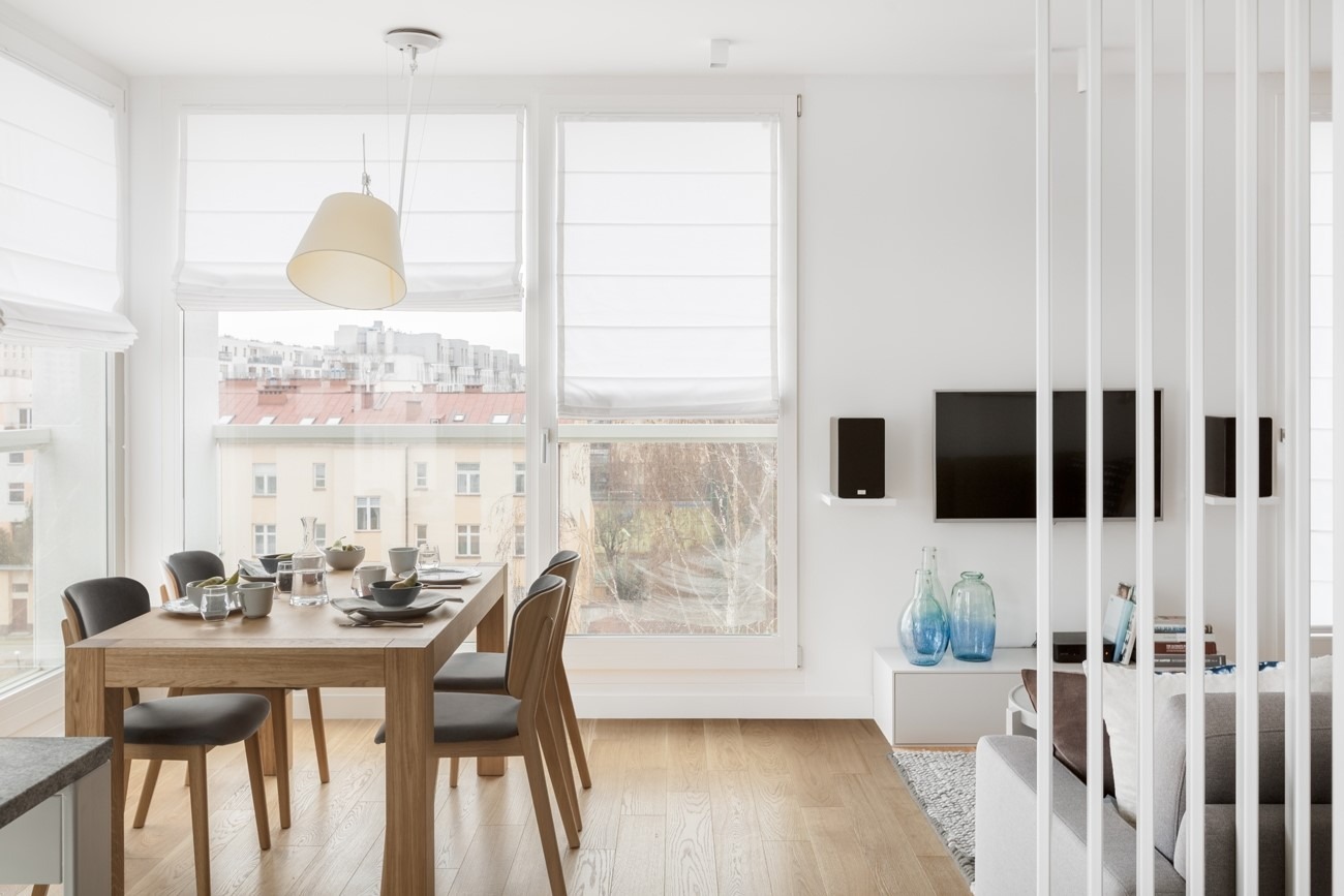 Jasne wnętrze jadalni z drewnianym stołem zastawionym naczyniami, szarymi krzesłami i białymi roletami rzymskimi na oknach, widok na budynki za oknem.