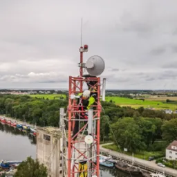 Technik w kasku i odblaskowej kamizelce wspina się po czerwonej wieży telekomunikacyjnej z antenami, w tle widok na rzekę, port, zieleń i turbiny wiatrowe pod pochmurnym niebem.