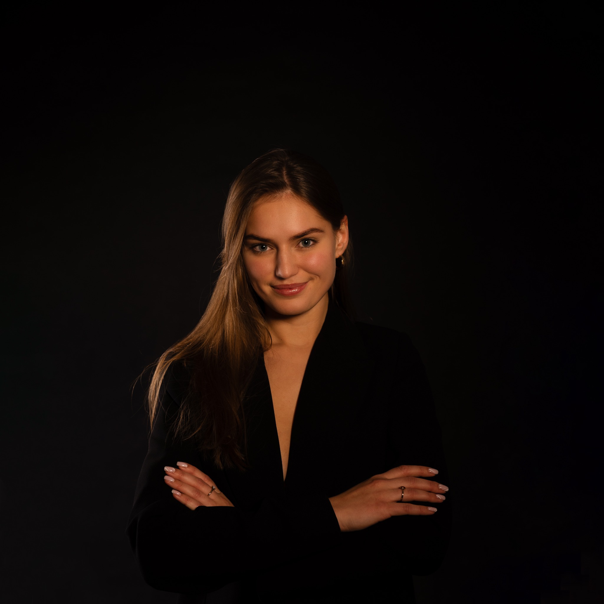 Portret uśmiechniętej kobiety w czarnym żakiecie na ciemnym tle, z rękami skrzyżowanymi na piersiach.