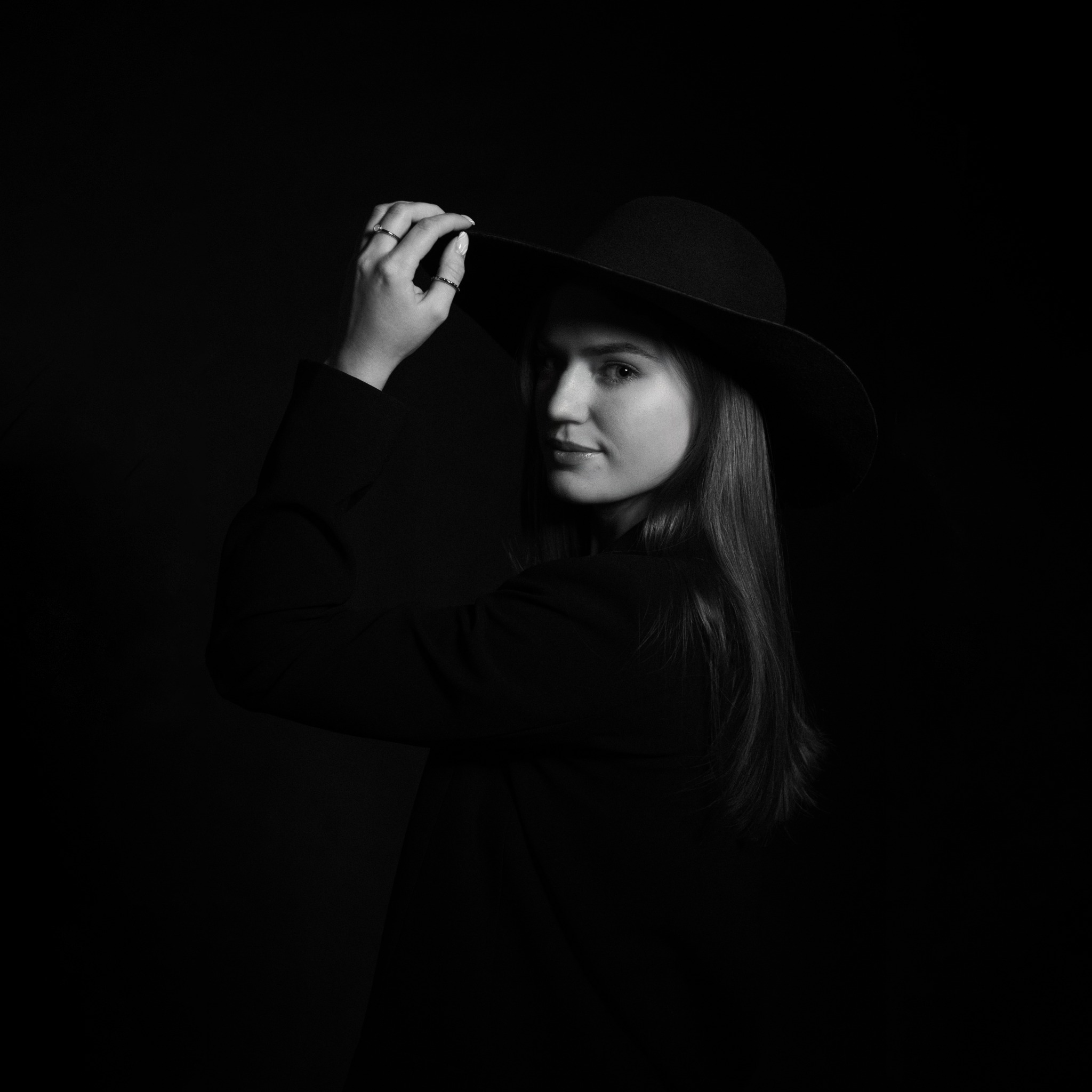 Portret kobiety w czarnym kapeluszu z szerokim rondem, w czerni i bieli, z delikatnym uśmiechem i dłonią dotykającą ronda kapelusza, na ciemnym tle.