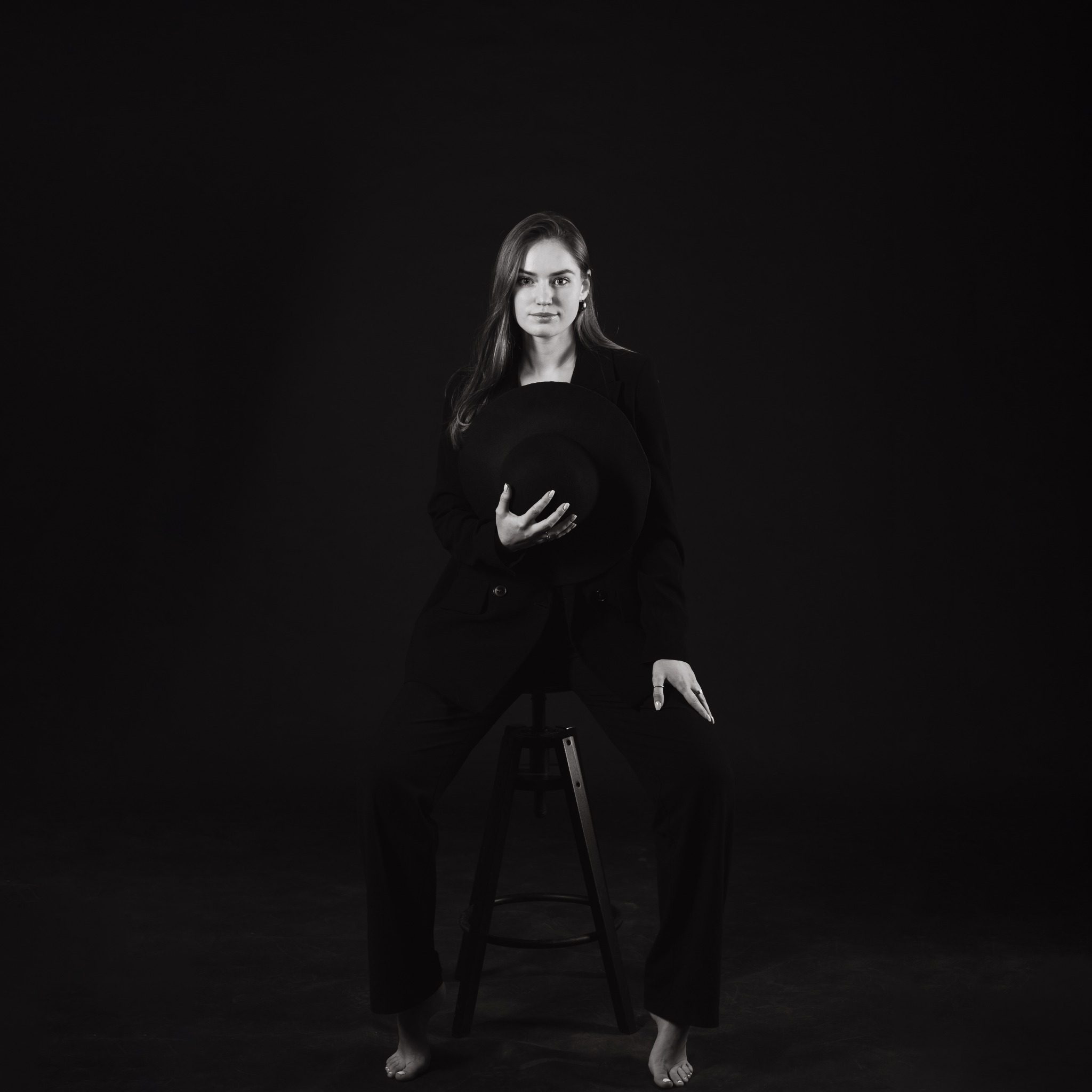 Portret kobiety w czarnym garniturze siedzącej na stołku, trzymającej czarny kapelusz, na czarnym tle, w stylu high fashion.