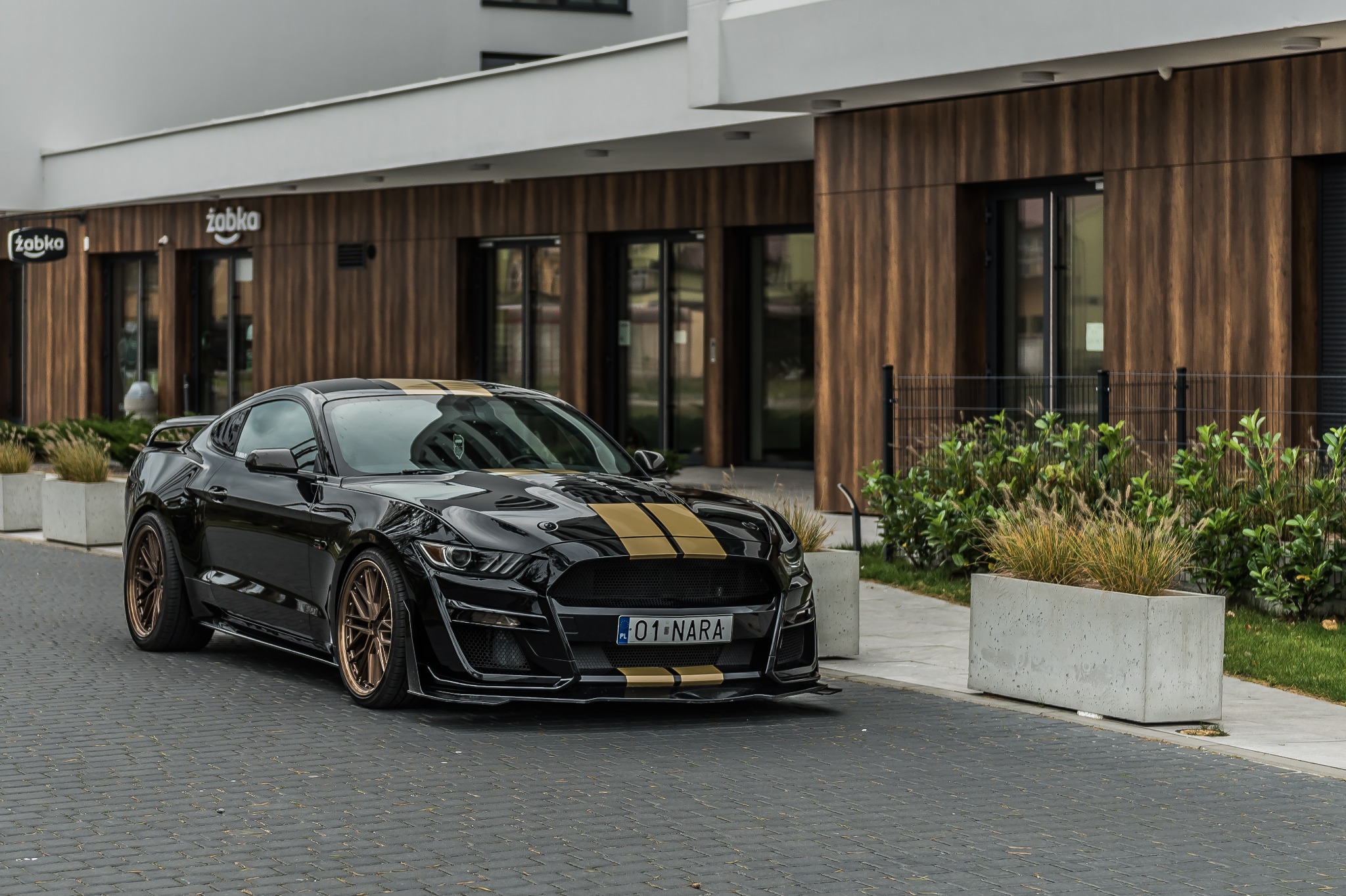 Czarny, sportowy samochód z złotymi pasami, zaparkowany na brukowanej drodze przed budynkiem z drewnianą elewacją i logiem Żabka.