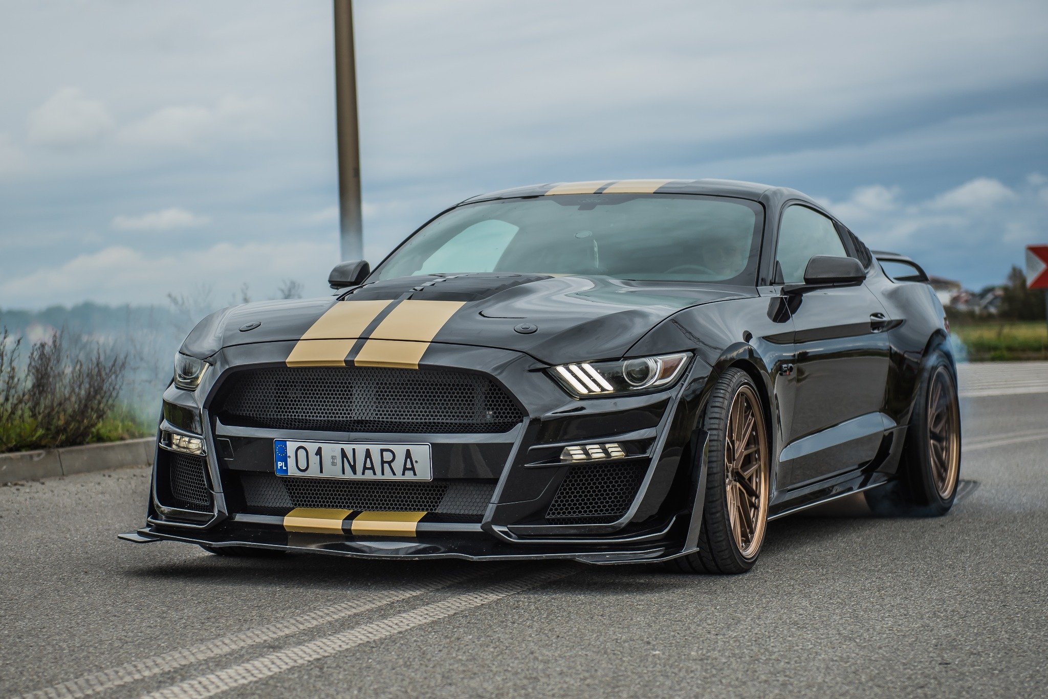 Czarny Ford Mustang z złotymi pasami na masce, tuningowane zawieszenie, dym z opon podczas jazdy.