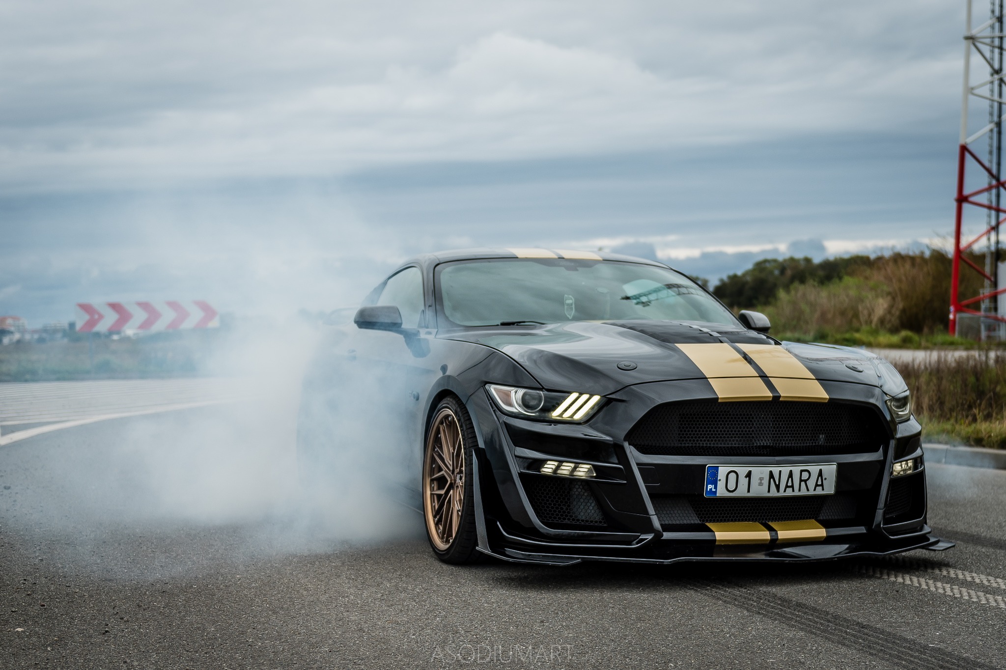 Czarny Mustang z dwoma złotymi pasami na masce, tablica rejestracyjna 01 NARA, dym spod kół podczas driftu, na tle pochmurnego nieba i wieży telekomunikacyjnej.