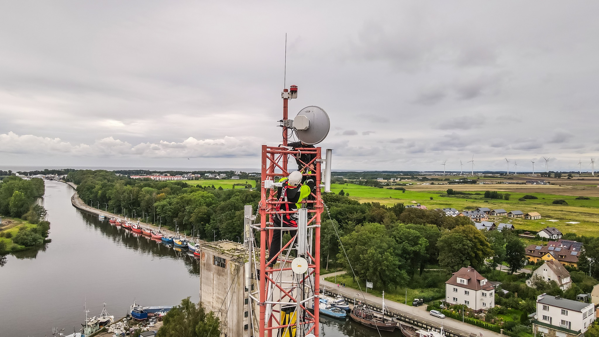 Technik w kasku i odblaskowej kamizelce wspina się po czerwonej wieży telekomunikacyjnej z antenami, w tle widok na rzekę, port, zieleń i turbiny wiatrowe pod pochmurnym niebem.