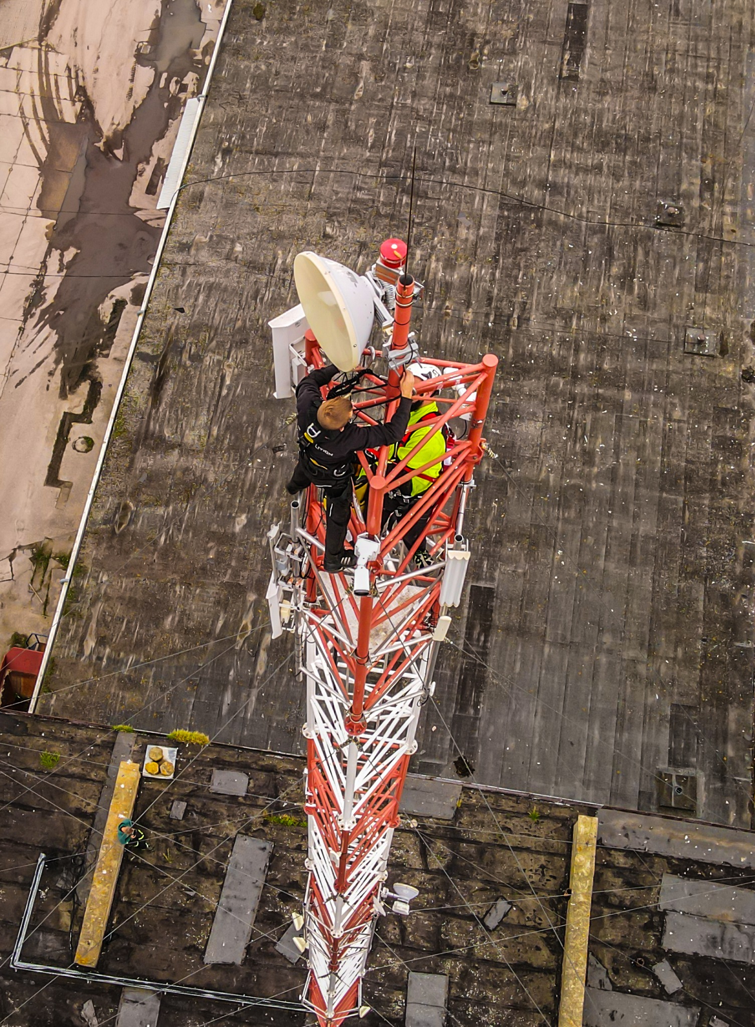 Pracownicy w kombinezonach zabezpieczających instalują antenę na szczycie wieży telekomunikacyjnej, widok z góry na tle dachu budynku.