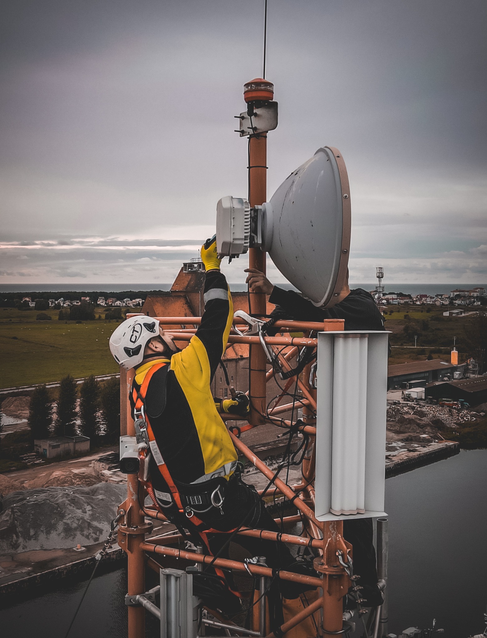 Dwóch techników w kaskach i uprzężach pracuje na pomarańczowej wieży telekomunikacyjnej z anteną paraboliczną, na tle pochmurnego nieba i oddalonego krajobrazu miejskiego z widokiem na morze.