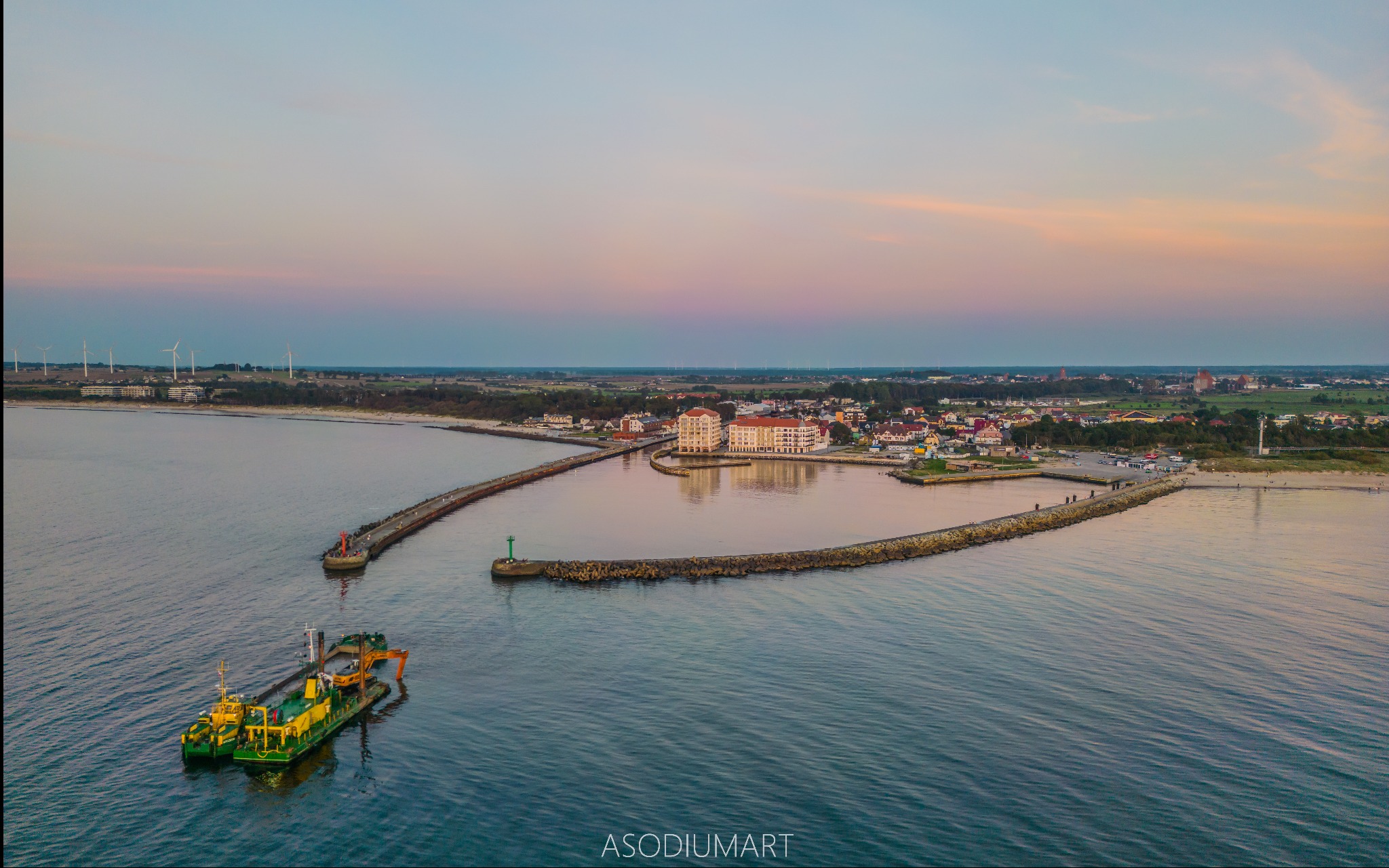 Widok z lotu ptaka na port z falochronami, miasto w tle i pogłębiarkę na pierwszym planie, oświetlone ciepłym światłem zachodzącego słońca.