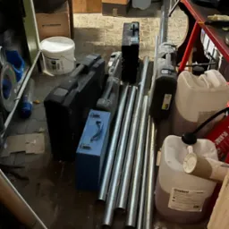 Stos rur instalacyjnych leżących na podłodze obok skrzynek narzędziowych i pojemników, w tle kartony i inne przedmioty w pomieszczeniu gospodarczym.