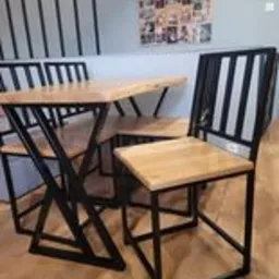 Stół i krzesła z metalowymi, czarnymi nogami o geometrycznym kształcie i drewnianymi blatami w jasnym odcieniu, ustawione na drewnianej podłodze w jasnym wnętrzu.