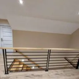 Nowoczesna balustrada wewnętrzna z czarnych, poziomych prętów i drewnianym pochwytem, zamontowana na antresoli z jasną podłogą i ścianami w odcieniach beżu. W tle widoczne drzwi z białym panelem.