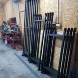 Elementy konstrukcyjne czarnych metalowych balustrad w warsztacie, widoczne spawy i przygotowane do montażu, w tle narzędzia i wyposażenie warsztatowe.