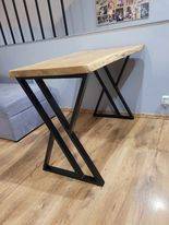 Stół z blatem z jasnego drewna o nieregularnych krawędziach, wsparty na czarnych, metalowych nogach w kształcie litery X. Ustawiony na drewnianej podłodze, na tle szarej ściany i częściowo...