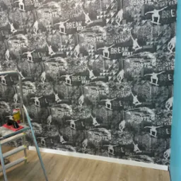 Ściana pokryta tapetą z motywem skateboardingu, drabina z narzędziami w trakcie pracy, widoczny fragment niebieskiej ściany obok.