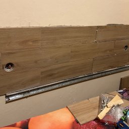 Krzysztof Czerny - Montaż płytek imitujących drewno na ścianie z widocznymi otworami na gniazdka elektryczne, listwa montażowa i narzędzia glazurnicze na pierwszym planie.