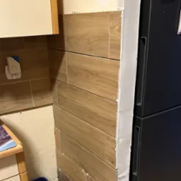 Układanie płytek imitujących drewno na ścianie w kuchni, widoczne krzyżyki dystansowe i niezagruntowana ściana obok lodówki