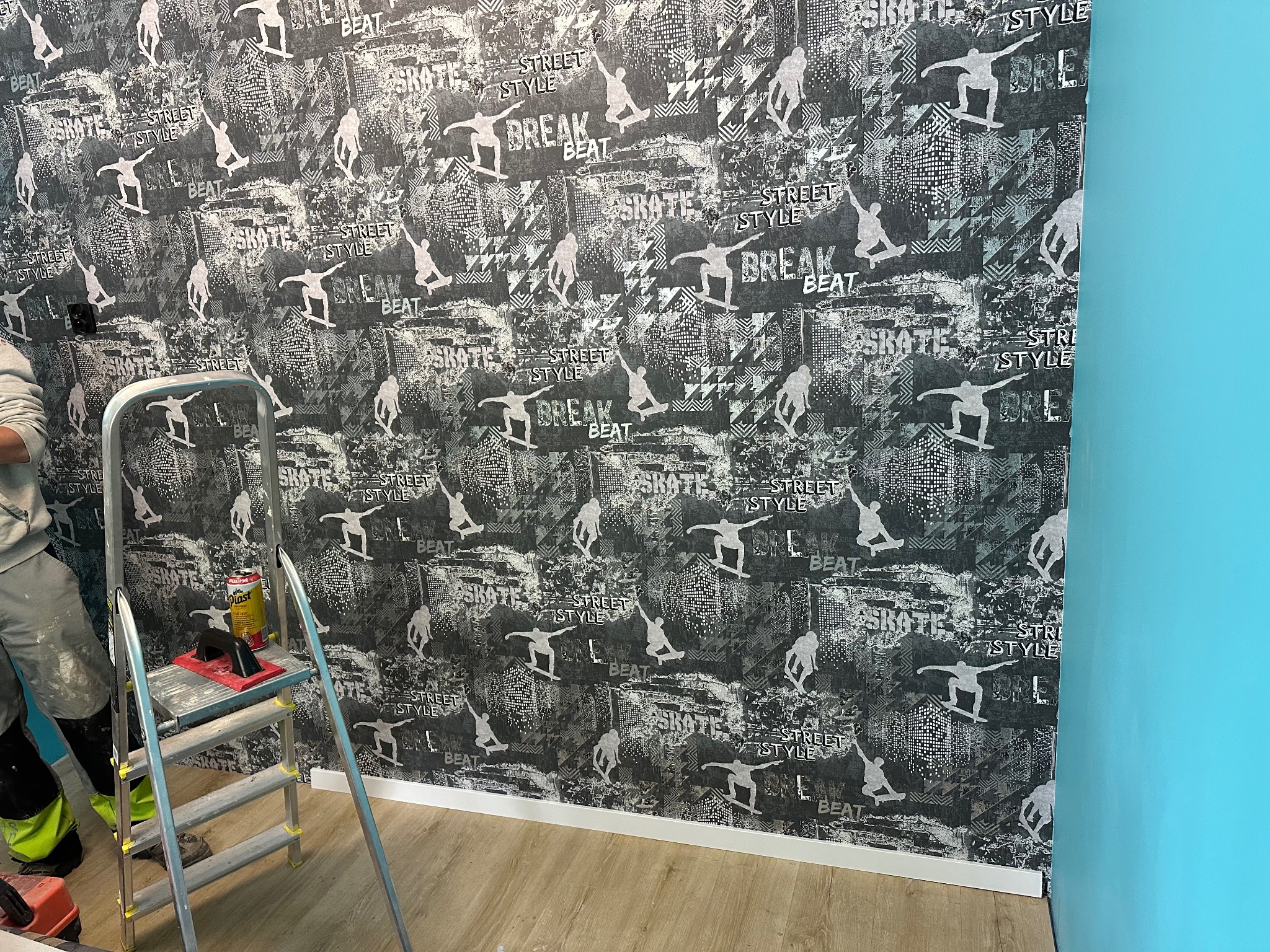 Ściana pokryta tapetą z motywem skateboardingu, drabina z narzędziami w trakcie pracy, widoczny fragment niebieskiej ściany obok.